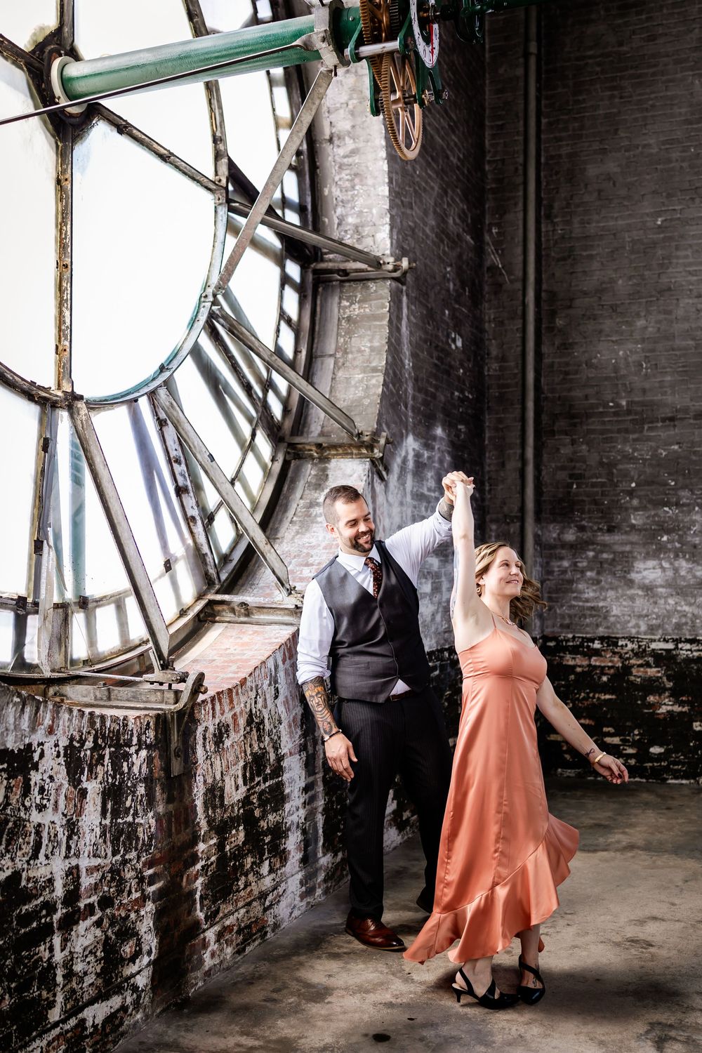 Dancing duo twirls gracefully in front of large clock windows in historic brick building.