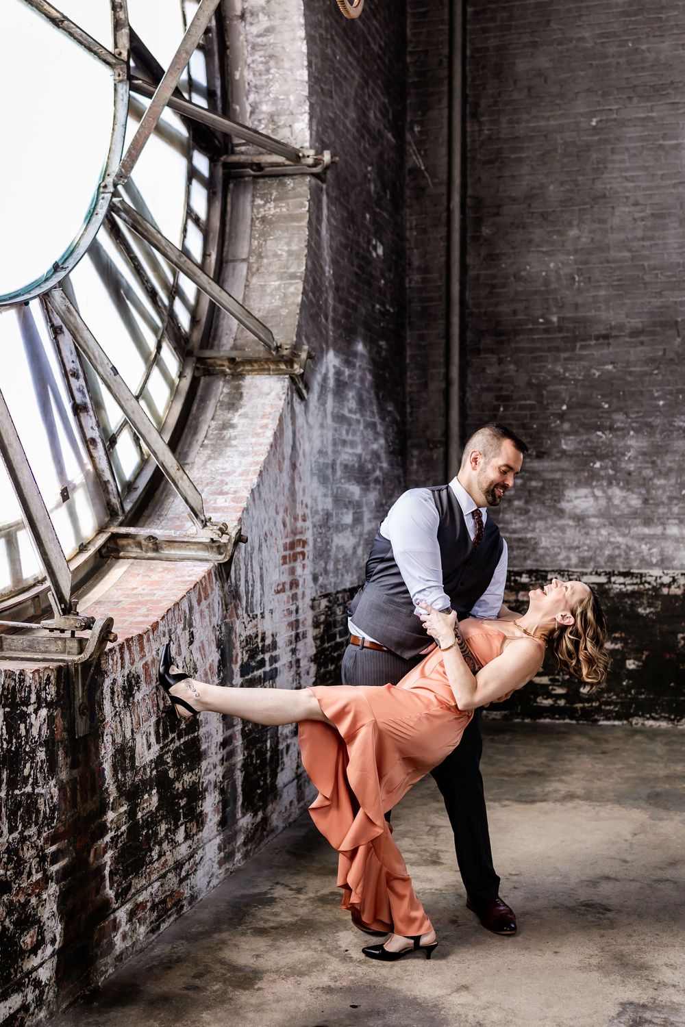 Romantic couple in formal attire dancing in an industrial space with brick walls and dramatic window lighting.