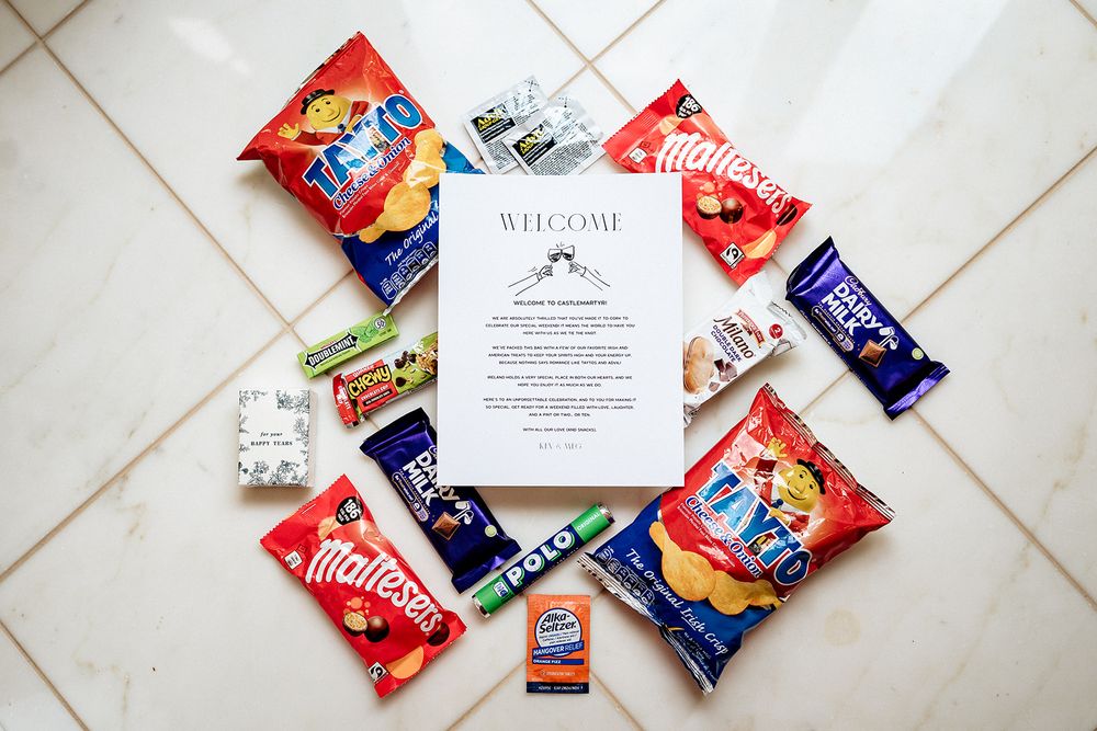 Various colorful snack bags and a wedding invitation arranged on white tile floor in a flat lay composition.