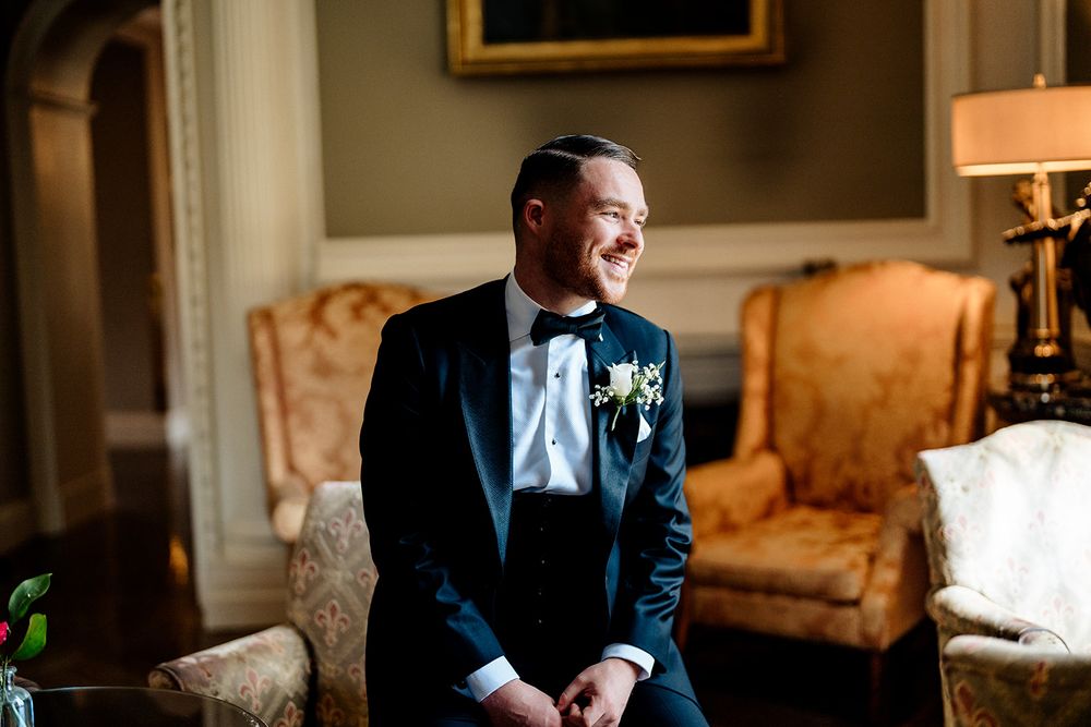 A groom in a tuxedo smiles while sitting at a wooden table in an elegantly decorated room.