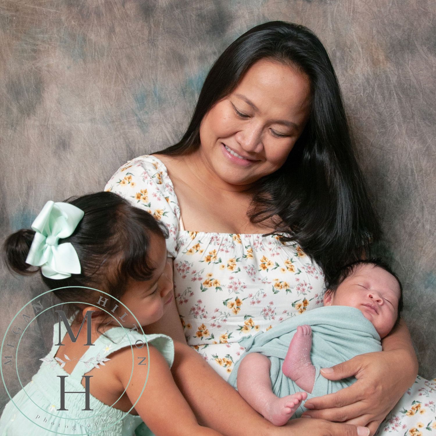 Mother in floral dress tenderly holds newborn baby in mint green swaddle while resting together on couch.