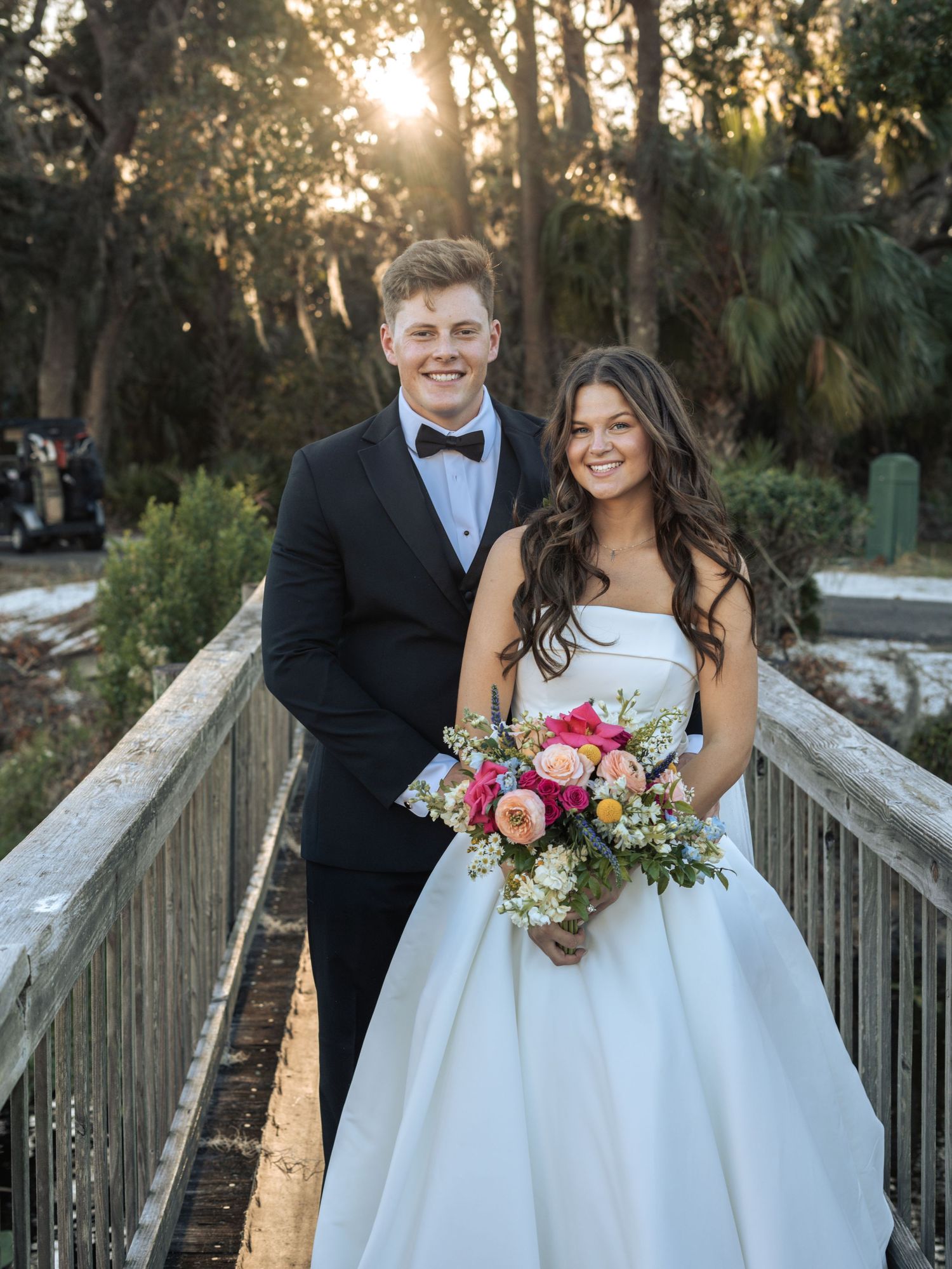 A newly married couple poses on a wooden bridge during sunset with the bride holding a vibrant pink bouquet.