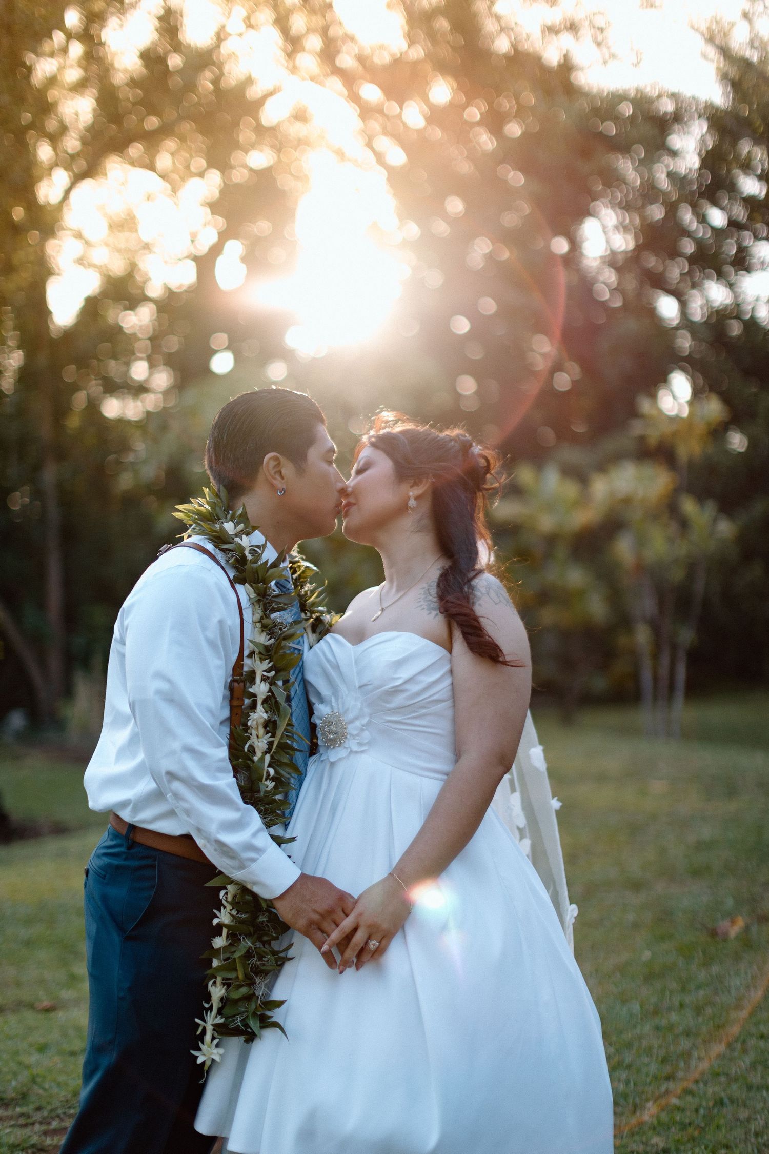 A wedding couple shares a romantic kiss at sunset while wearing traditional Hawaiian leis in an outdoor garden setting.
