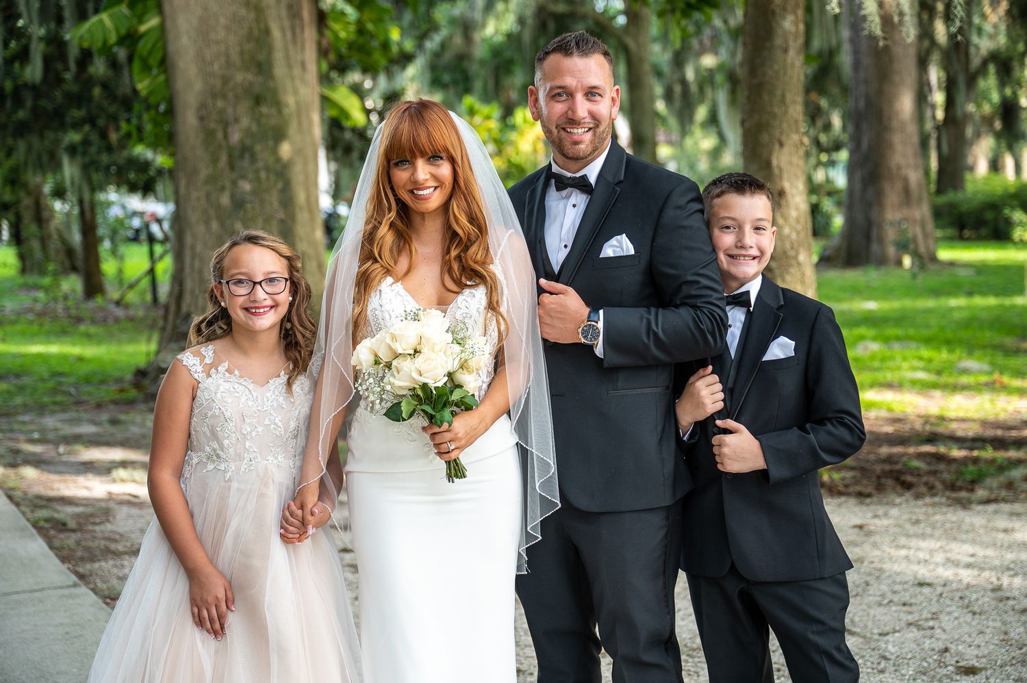 Happy wedding party stands together on a garden path surrounded by green trees and holding white floral bouquet.