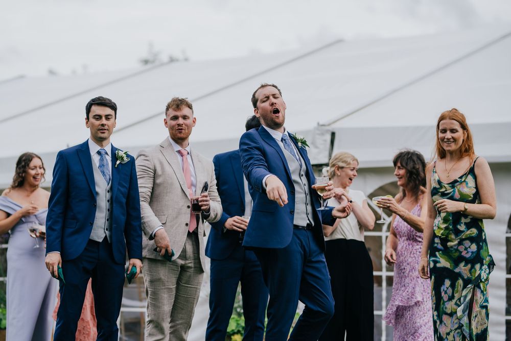 Wedding guests on a grassy lawn raise drinks in celebratory toasts at an outdoor reception.