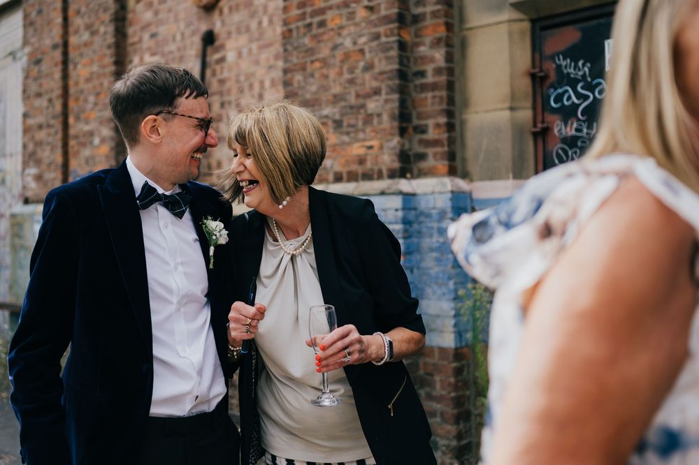 A happy couple celebrates with wine and laughter surrounded by wedding guests against a brick wall.
