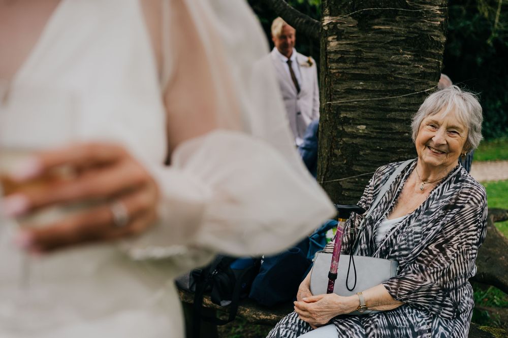 An elderly woman in a patterned dress smiles joyfully while seated at an outdoor wedding celebration.
