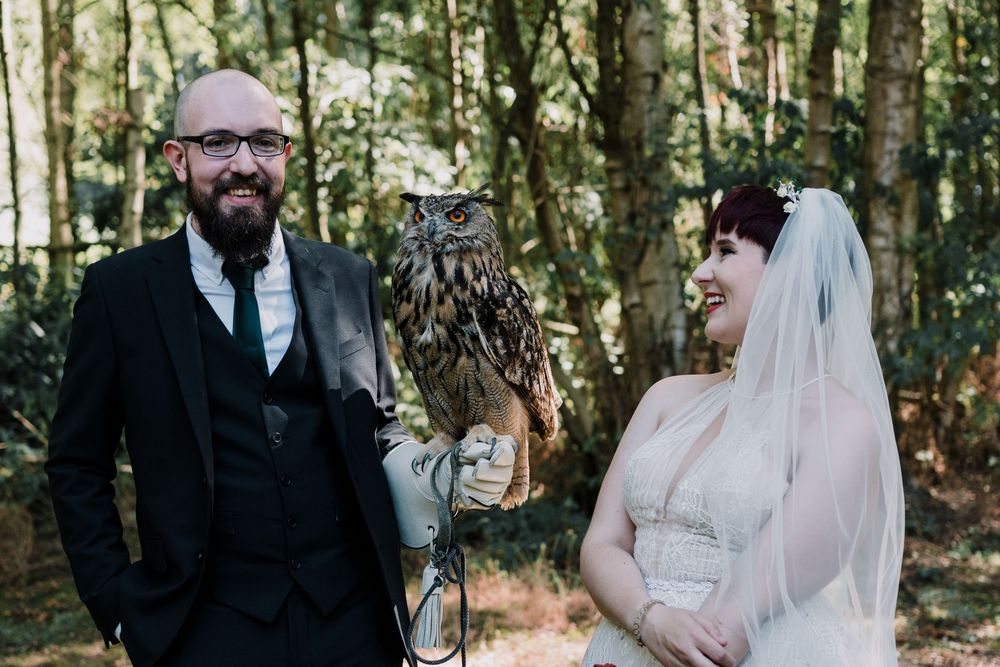 A wedding couple poses in a forest setting with a large owl perched between them during their outdoor ceremony.