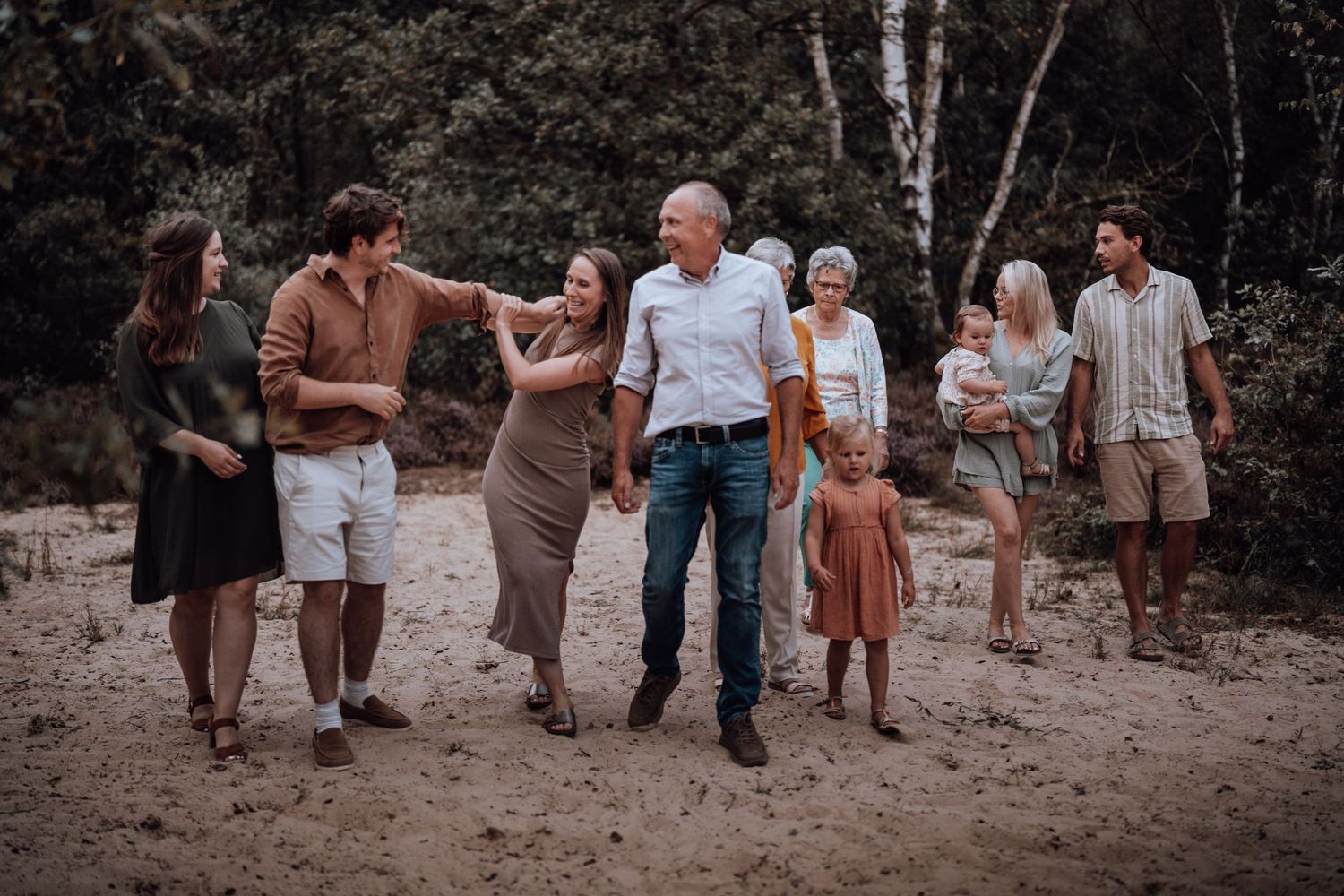 Een groep mensen in vrijetijdskleding staat bij elkaar in een rustieke setting buiten, met dennenbomen in de achtergrond.