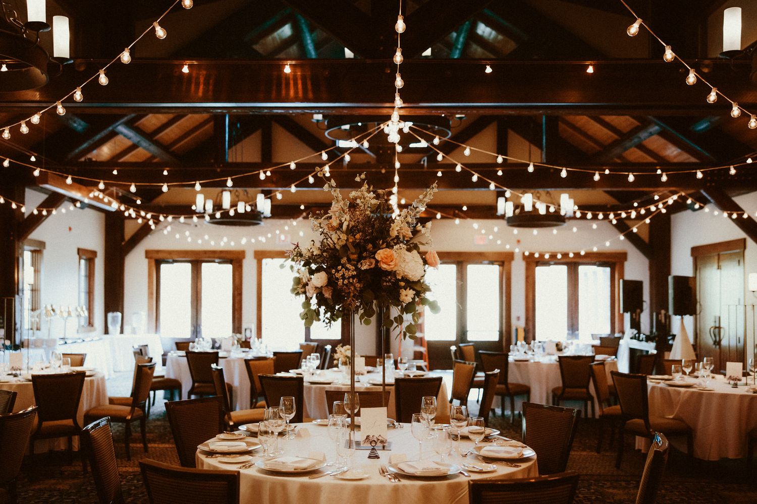 Elegant wedding reception with floral centrepiece, round tables, and string lights in a rustic venue with wooden beams and warm ambiance.