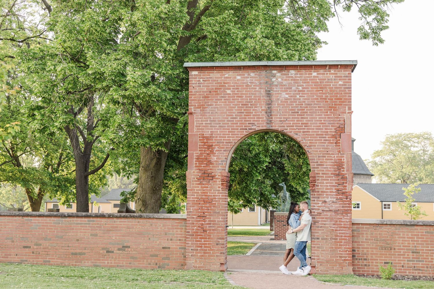 A couple embraces beneath a historic brick archway on a college campus surrounded by green trees.