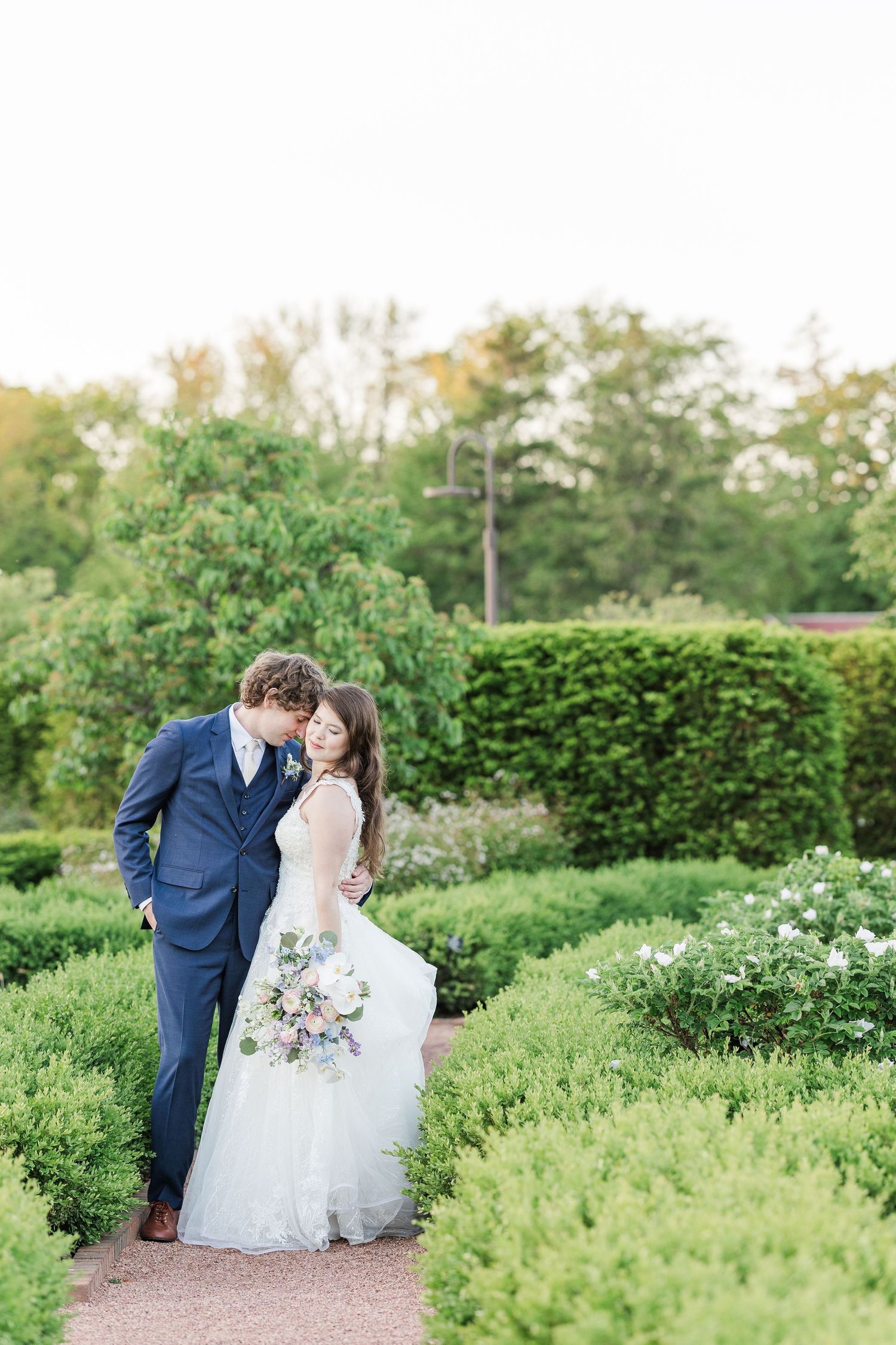 A wedding couple kisses on a garden path surrounded by lush greenery and hedges on a spring day.