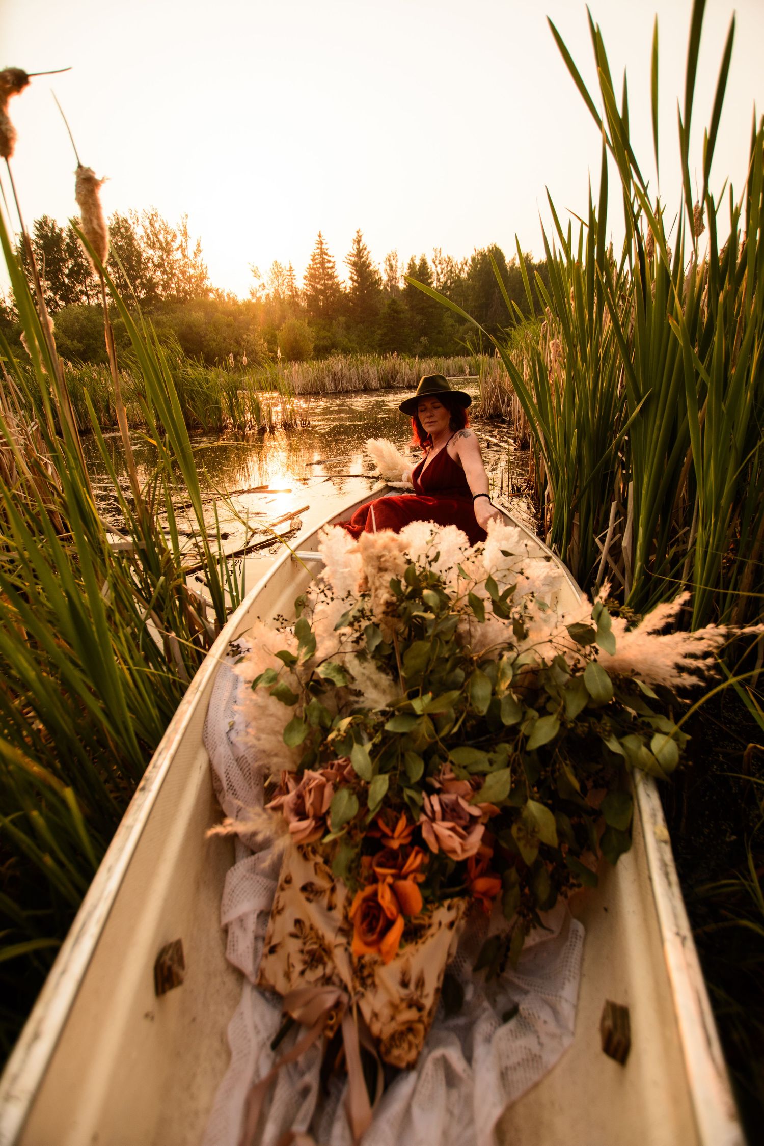 Boat filled with flowers and greens sailing through calm waters at sunset with cattails along the shore.