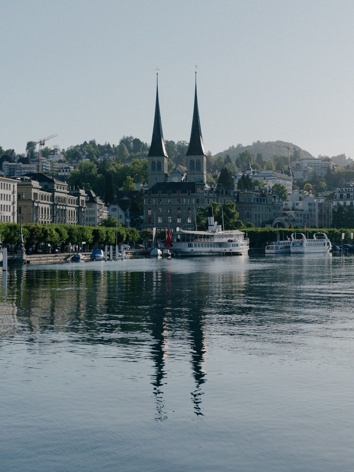 Historic church spires and buildings reflect in the calm waters of Lake Lucerne with boats docked along the shoreline.