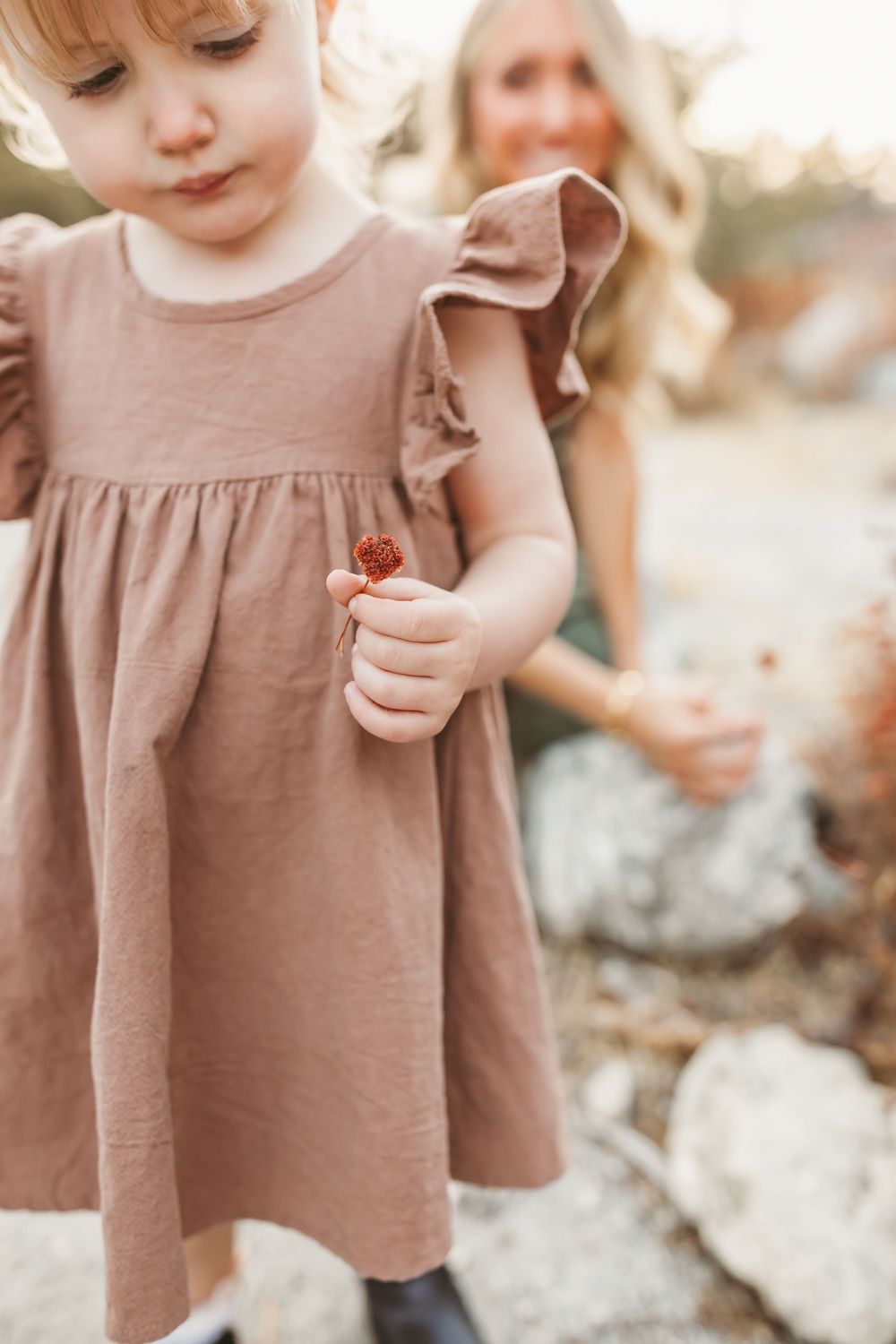 A toddler in a dusty pink dress with ruffled sleeves holds something small in their hand while outdoors.