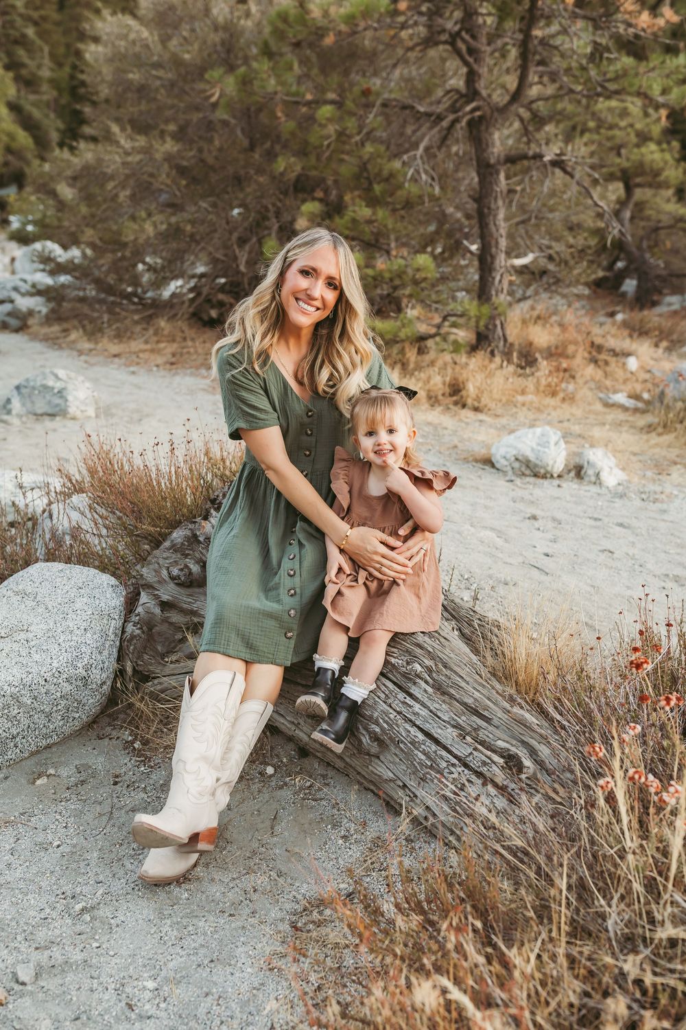 A parent and child in coordinating outfits sit together on a fallen log in a rugged outdoor setting.