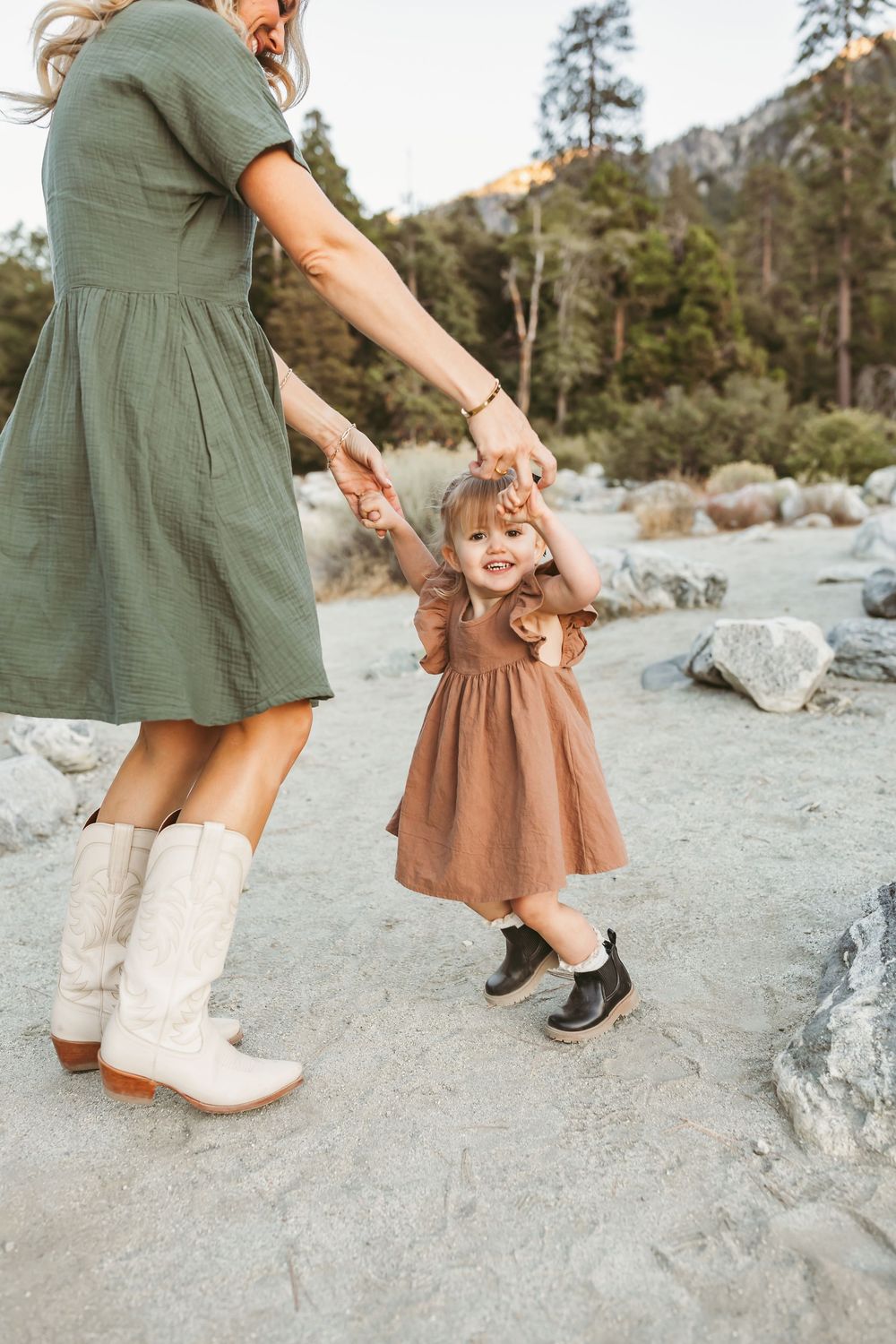 An adult wearing a green dress and boots holds hands with a toddler in a brown dress dancing on a rocky beach.