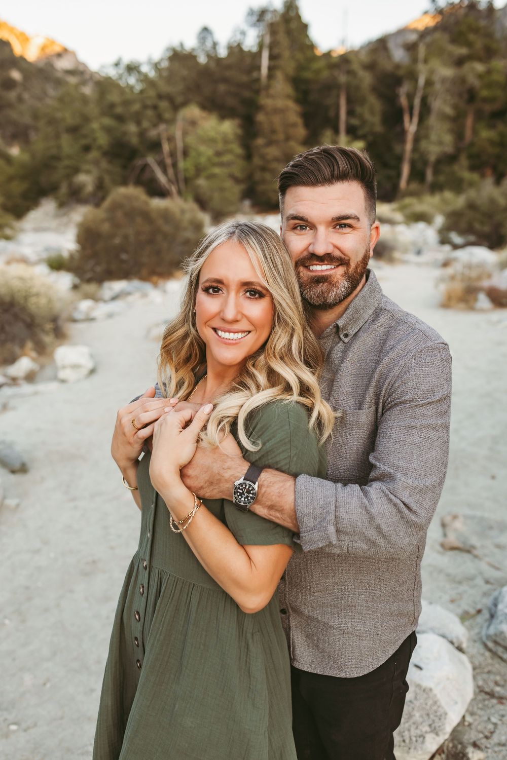A couple poses together outdoors in a desert landscape at sunset wearing grey and olive green casual attire.