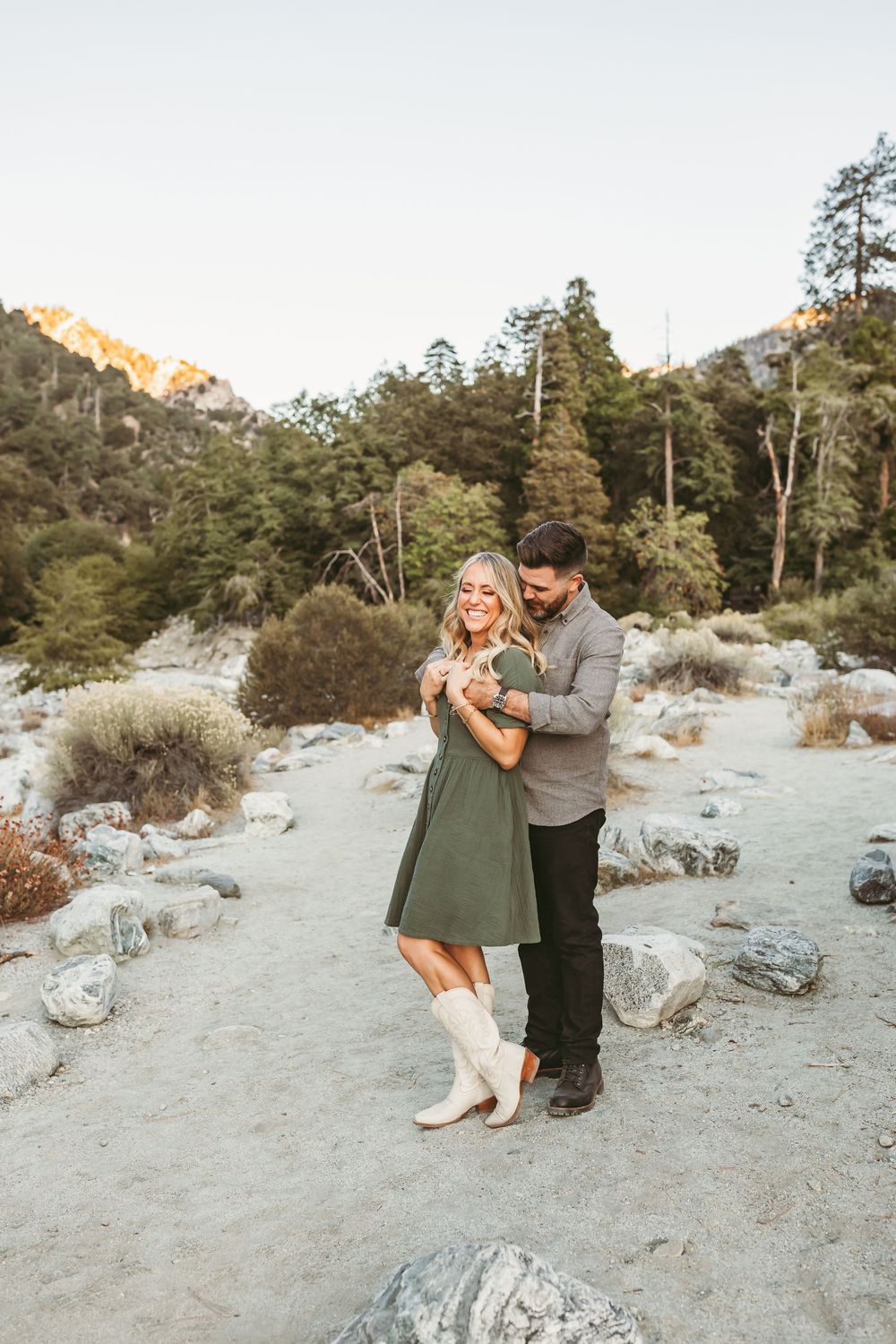 A couple embraces and dances together against a scenic mountain backdrop with pine trees and rocks.