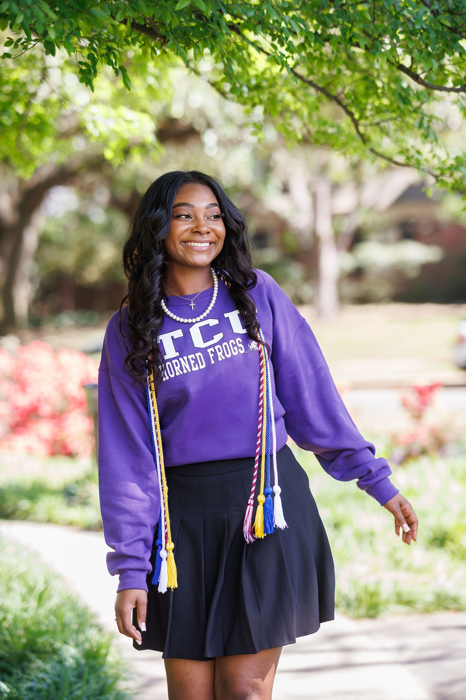 A young woman in a purple TCU college sweatshirt and black skirt poses outdoors with graduation cords on a sunny day in Dallas
