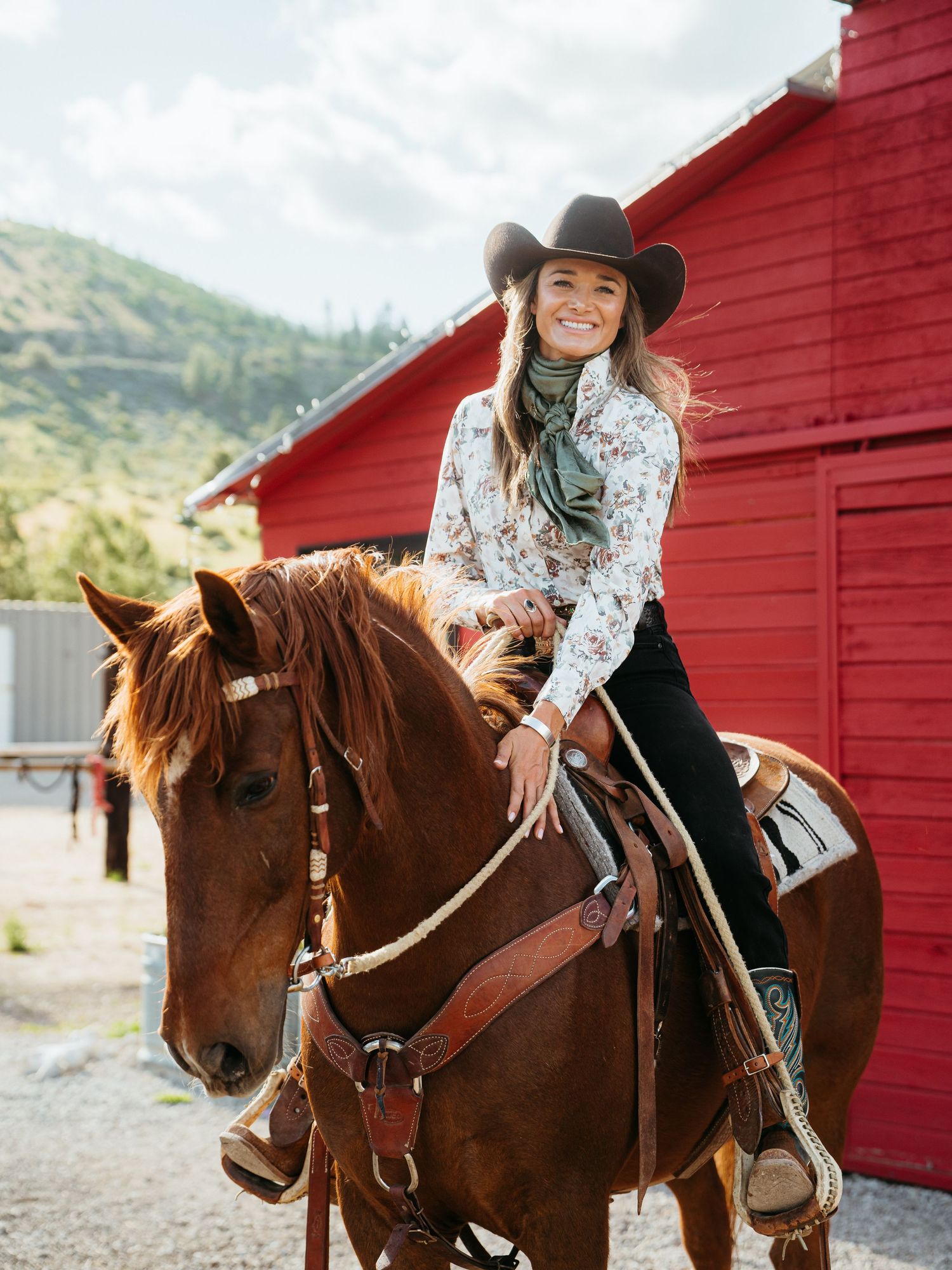 A cowgirl in a white hat and denim lovingly pets a light-colored horse at a ranch corral on a sunny day.
