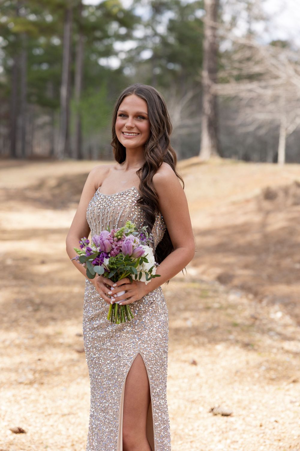 A person in a sparkly silver formal gown with a slit holds a purple and white floral bouquet outdoors.