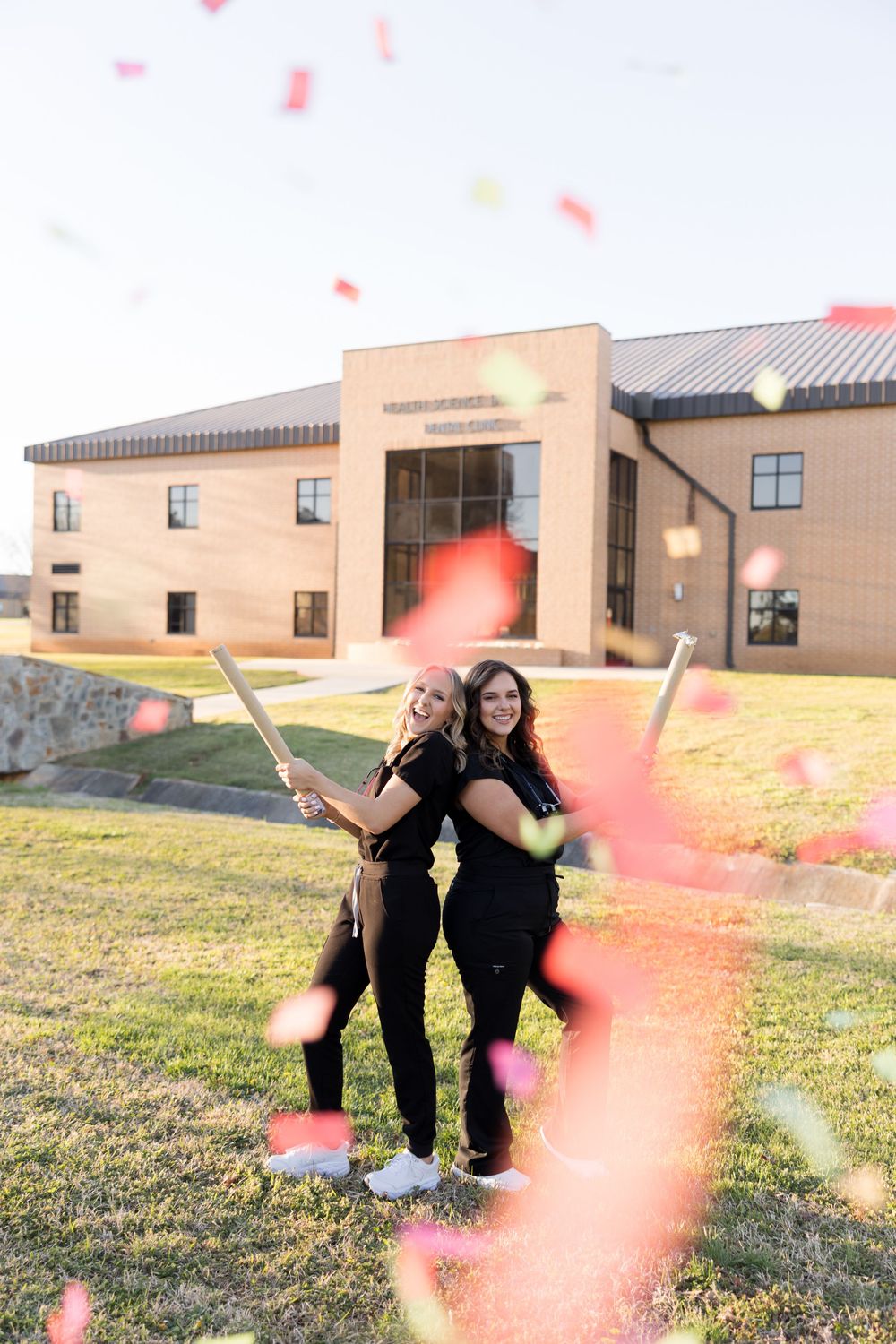 Two people in black outfits stand back-to-back holding sparklers outside a beige building on a grassy lawn at sunset.