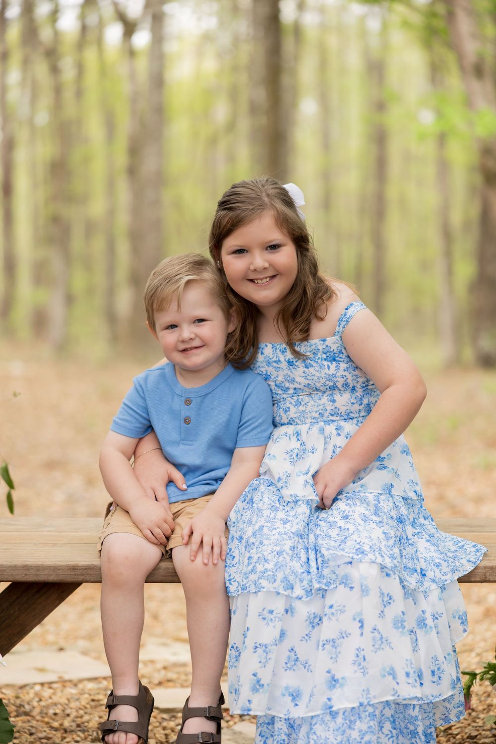 A pair of children sit together on a wooden bench in a sunlit forest with blurred trees in the background.