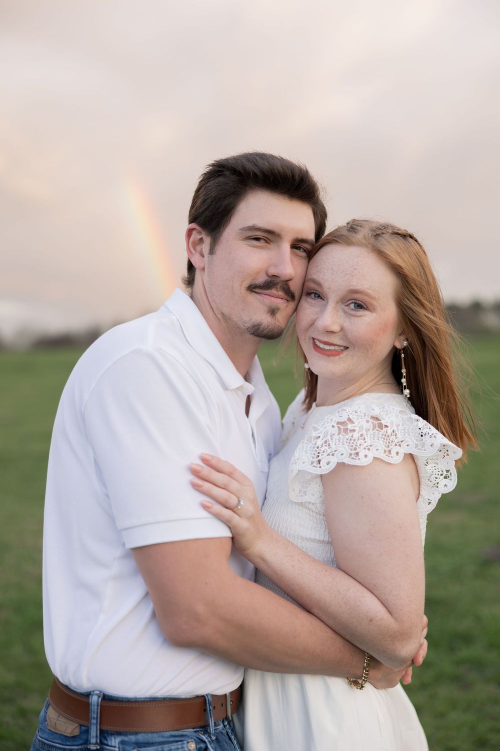 A couple embraces in a field at sunset while a rainbow appears in the cloudy sky behind them.