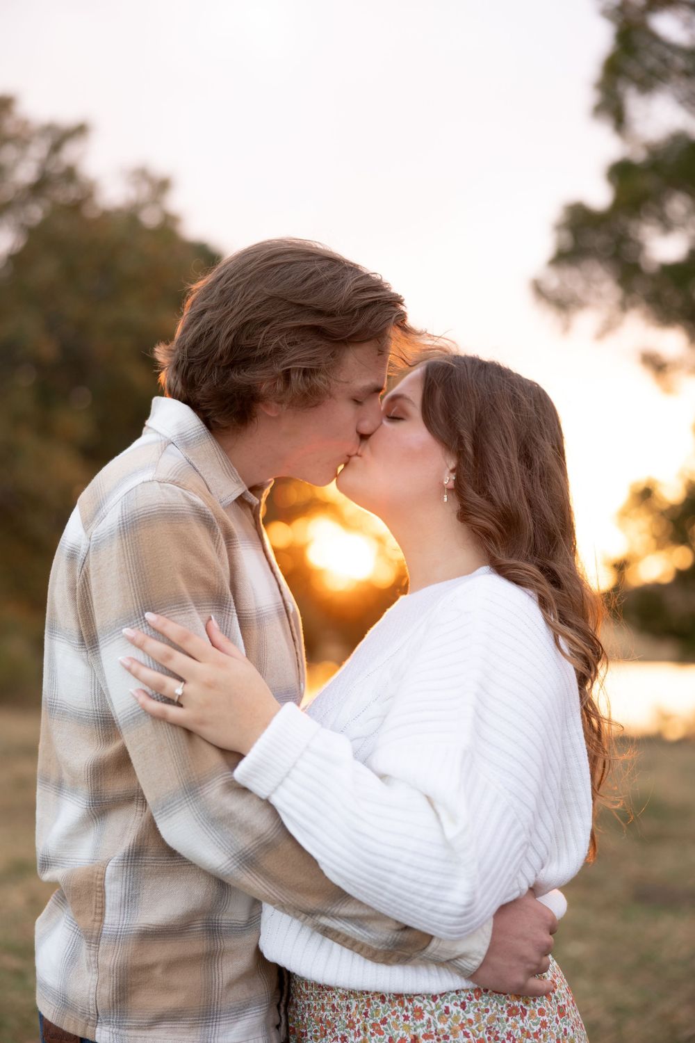 A romantic couple shares a tender kiss at sunset while embracing outdoors in warm golden light.