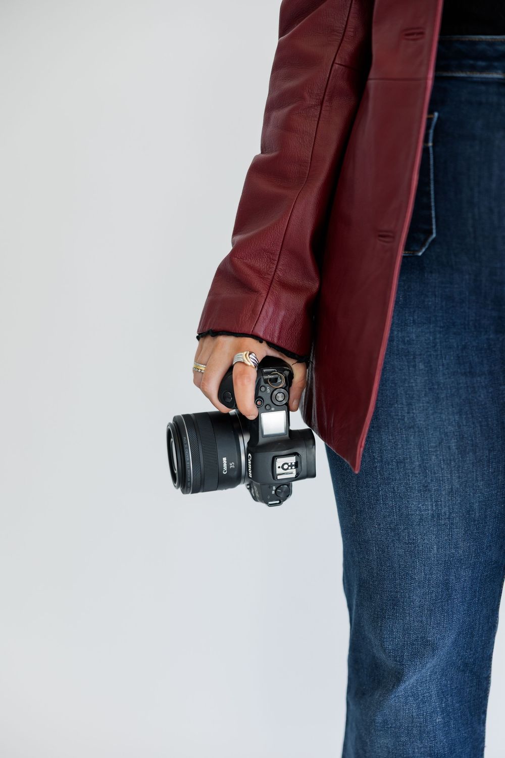 Someone wearing a burgundy jacket and jeans holds a black camera with lens against white background.