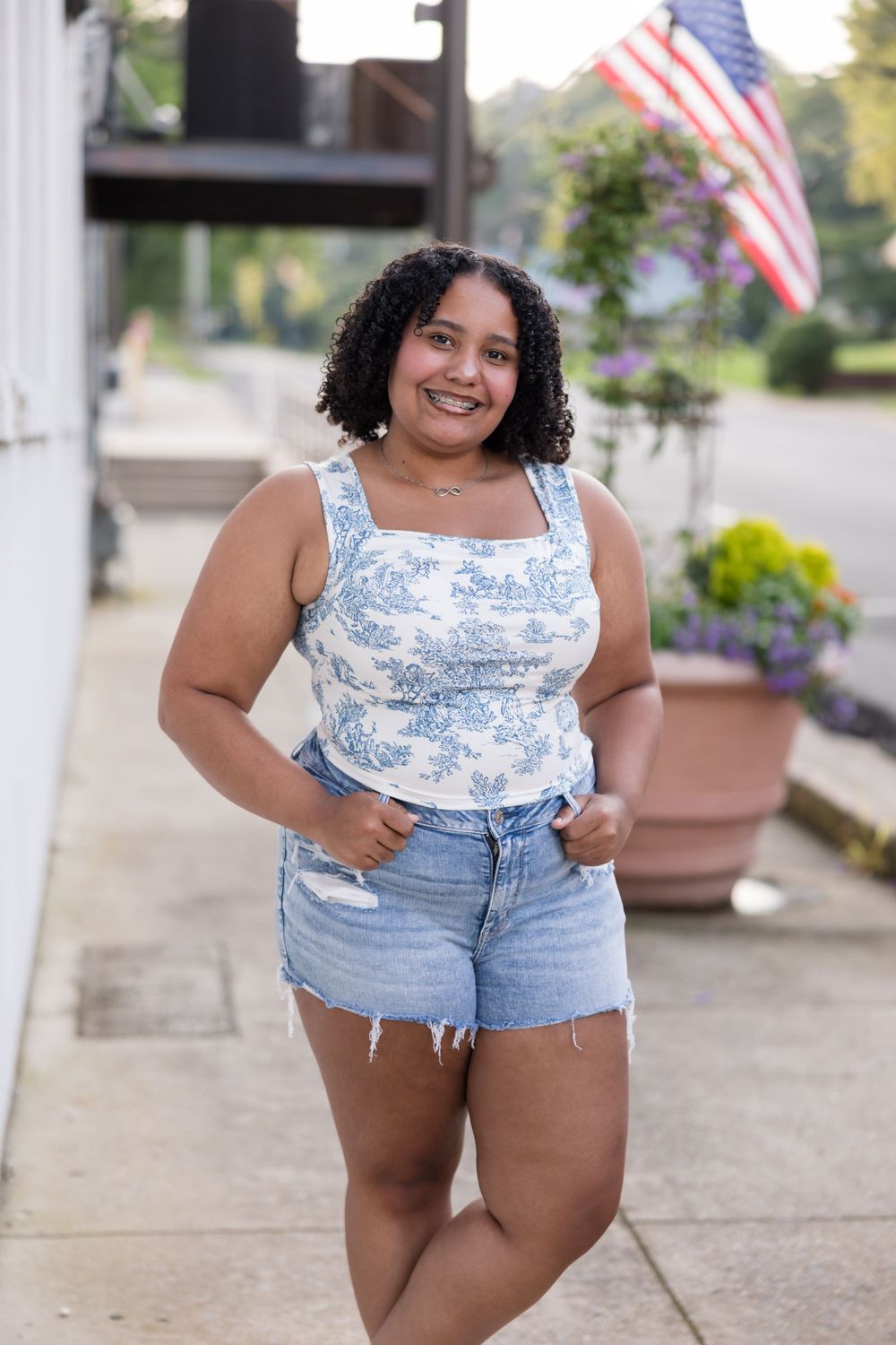 A figure in a blue and white toile-print tank top and denim shorts poses outdoors with American flags in the background.