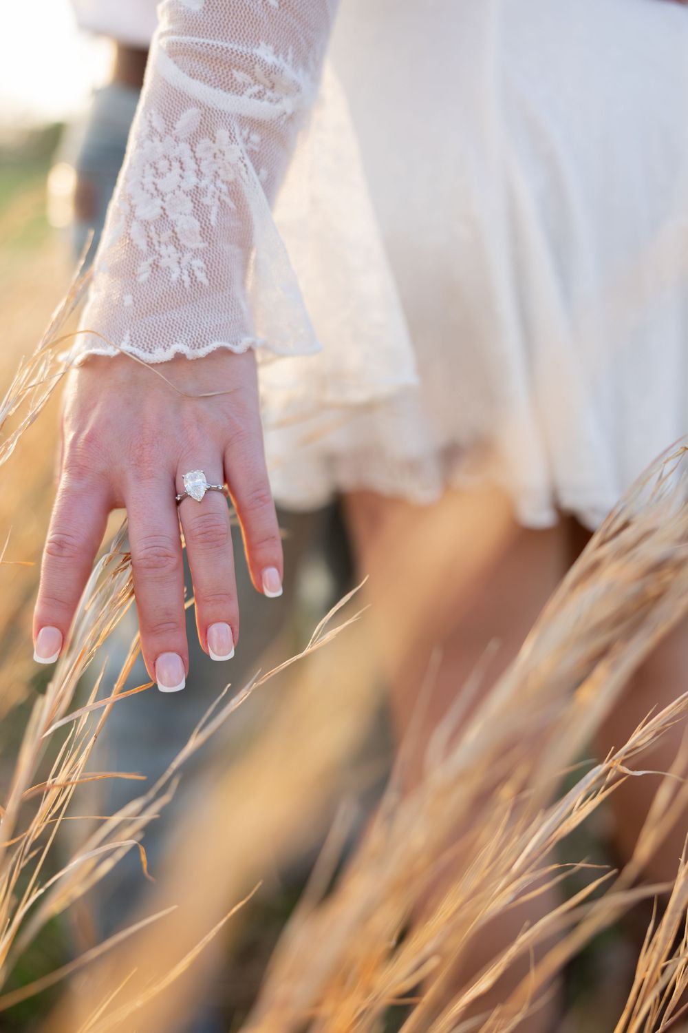 Close-up of delicate lace-sleeved hand wearing an engagement ring brushing against golden wheat stalks in sunlight.