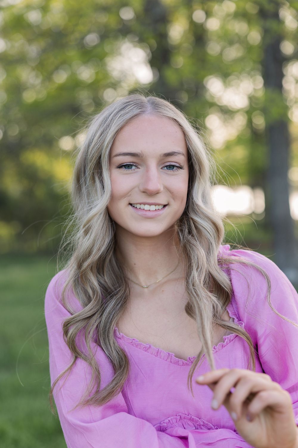 A person wearing a pink top poses outdoors with wavy blonde hair in a natural setting with bokeh background.