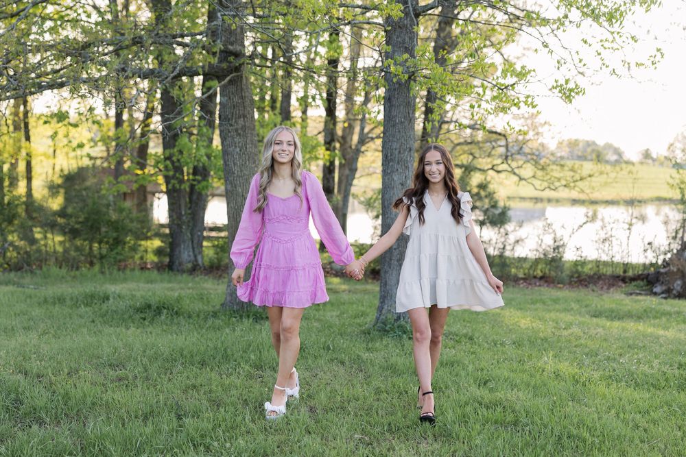 Two people hold hands while walking together in flowy dresses through a scenic grassy area near trees and water.