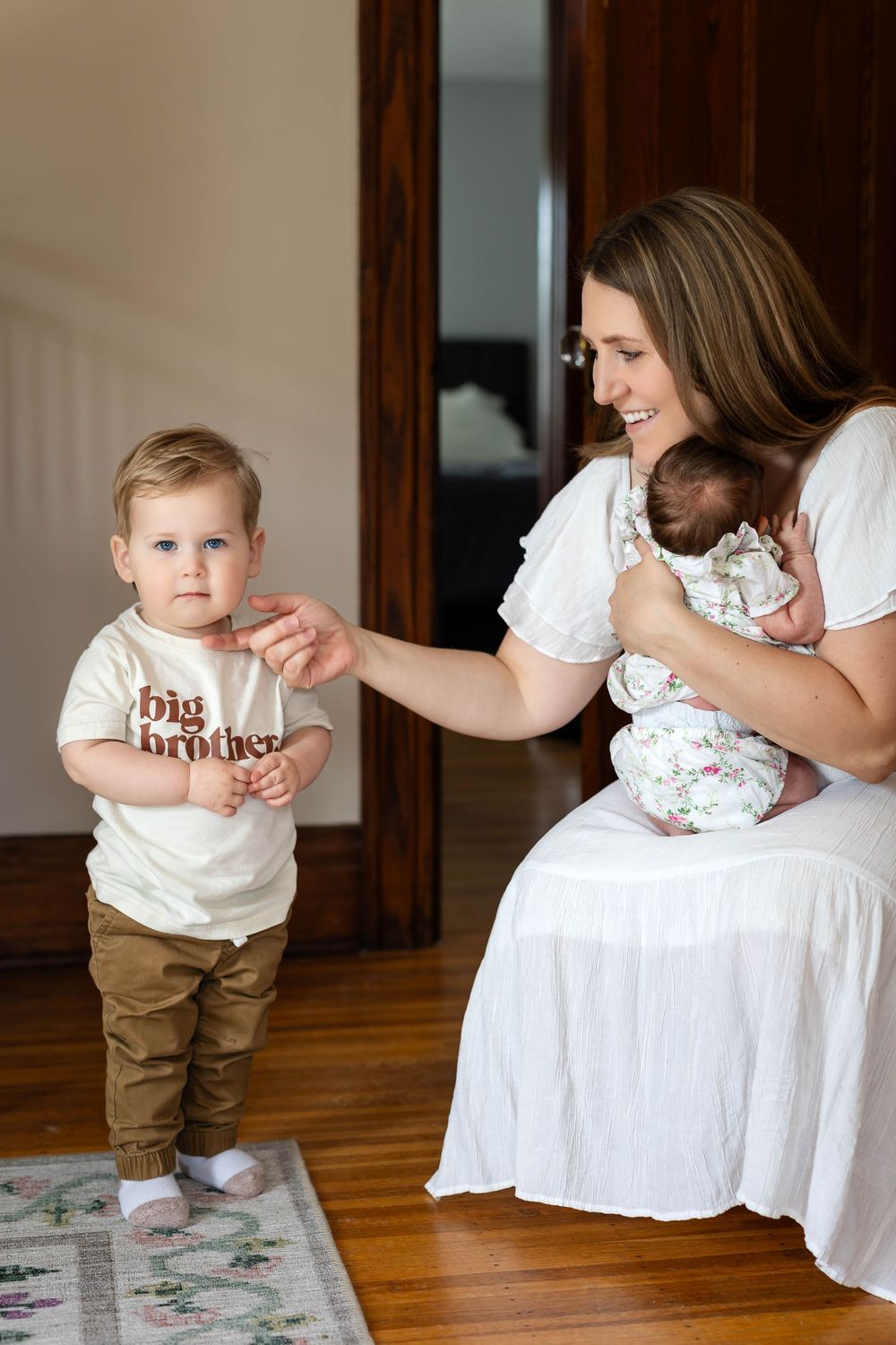 Mother in white dress sits with newborn while toddler in 'big brother' shirt stands nearby.