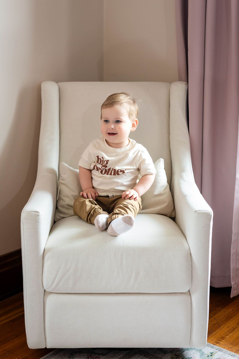 Toddler sitting happily in a white armchair wearing a white shirt and brown pants.