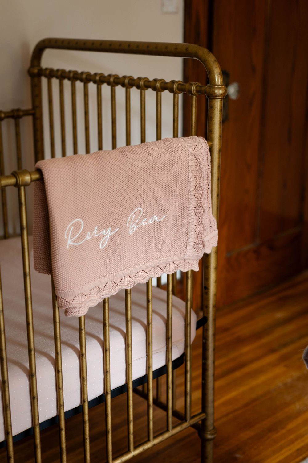 Pink baby blanket with embroidered name draped over vintage brass crib railing.