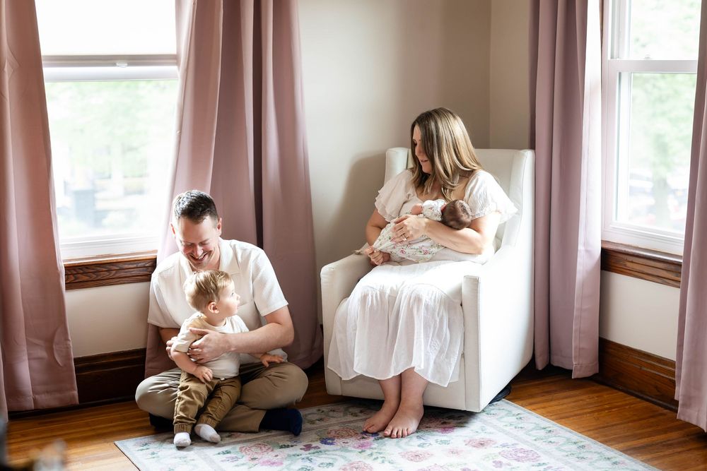 Family sits together in a cozy room with pink curtains as they hold their newborn baby.