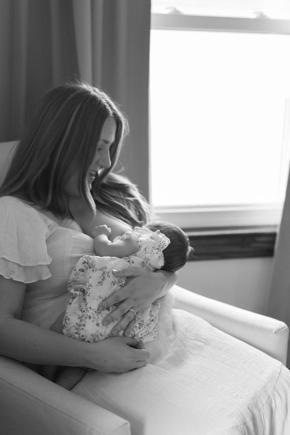 Mother in white dress lovingly cradles newborn baby in a sunlit room.