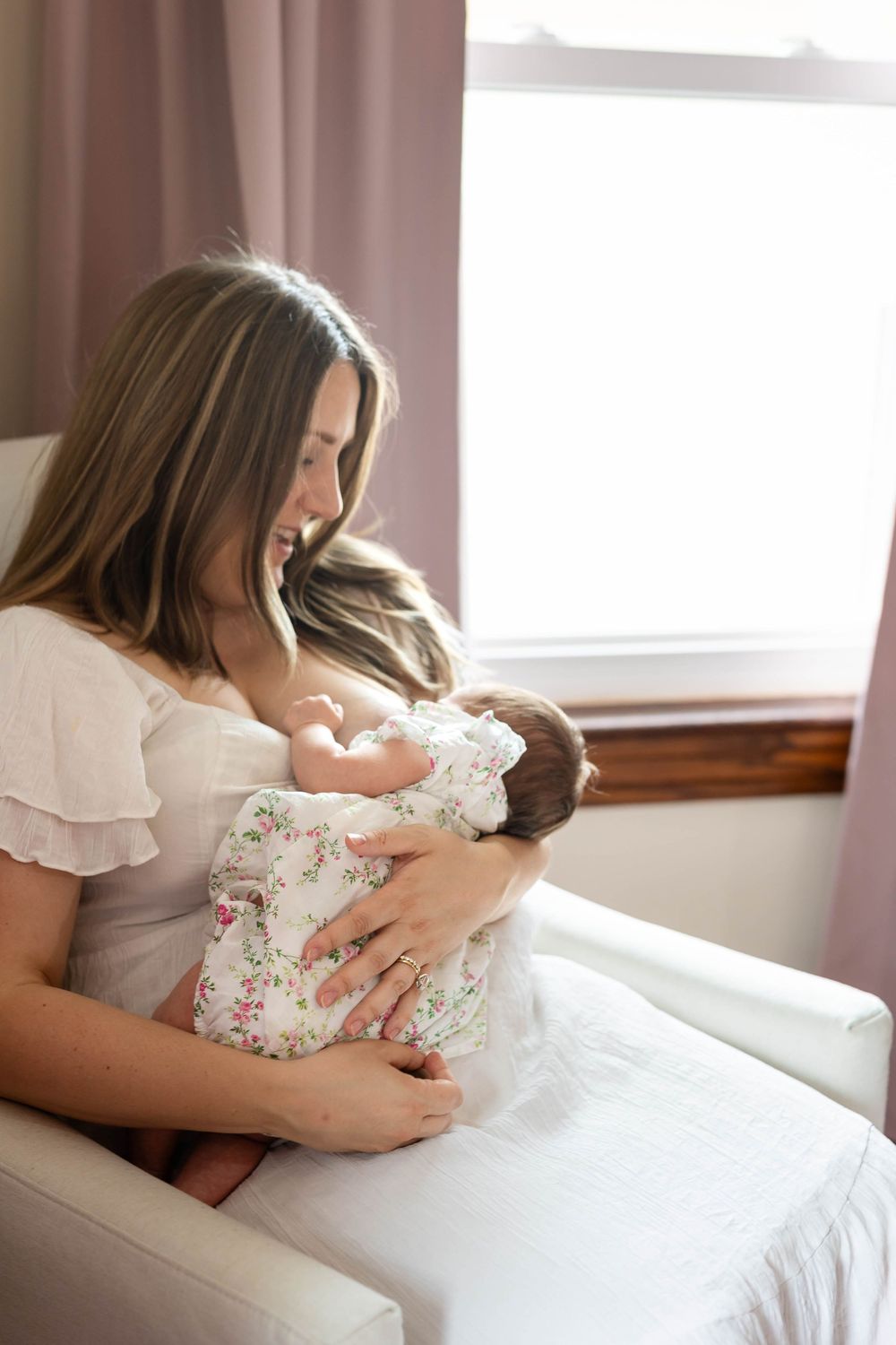 A woman in a white ruffled dress sits by a window while lovingly holding a sleeping infant.