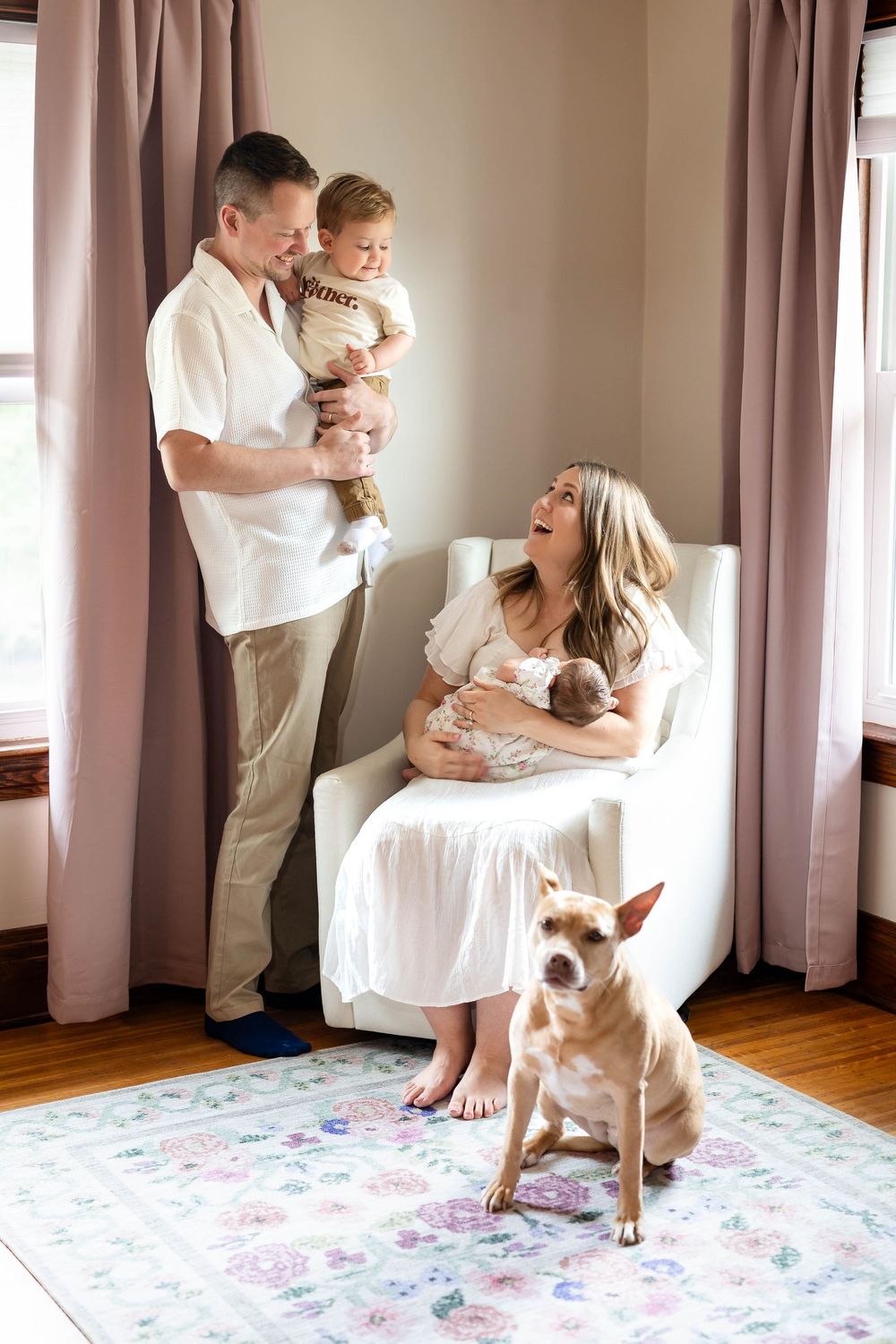 Family portrait showing parents with newborn baby and their dog on a floral rug by pink curtains.