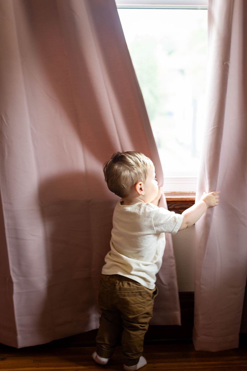 Young child reaching up to touch pink curtains by a window.