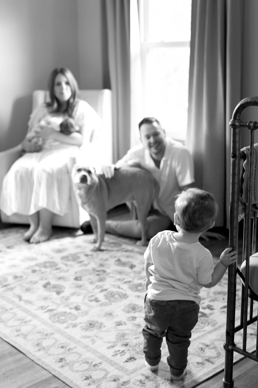 Black and white family photo in nursery with baby, dog, and vintage metal crib.