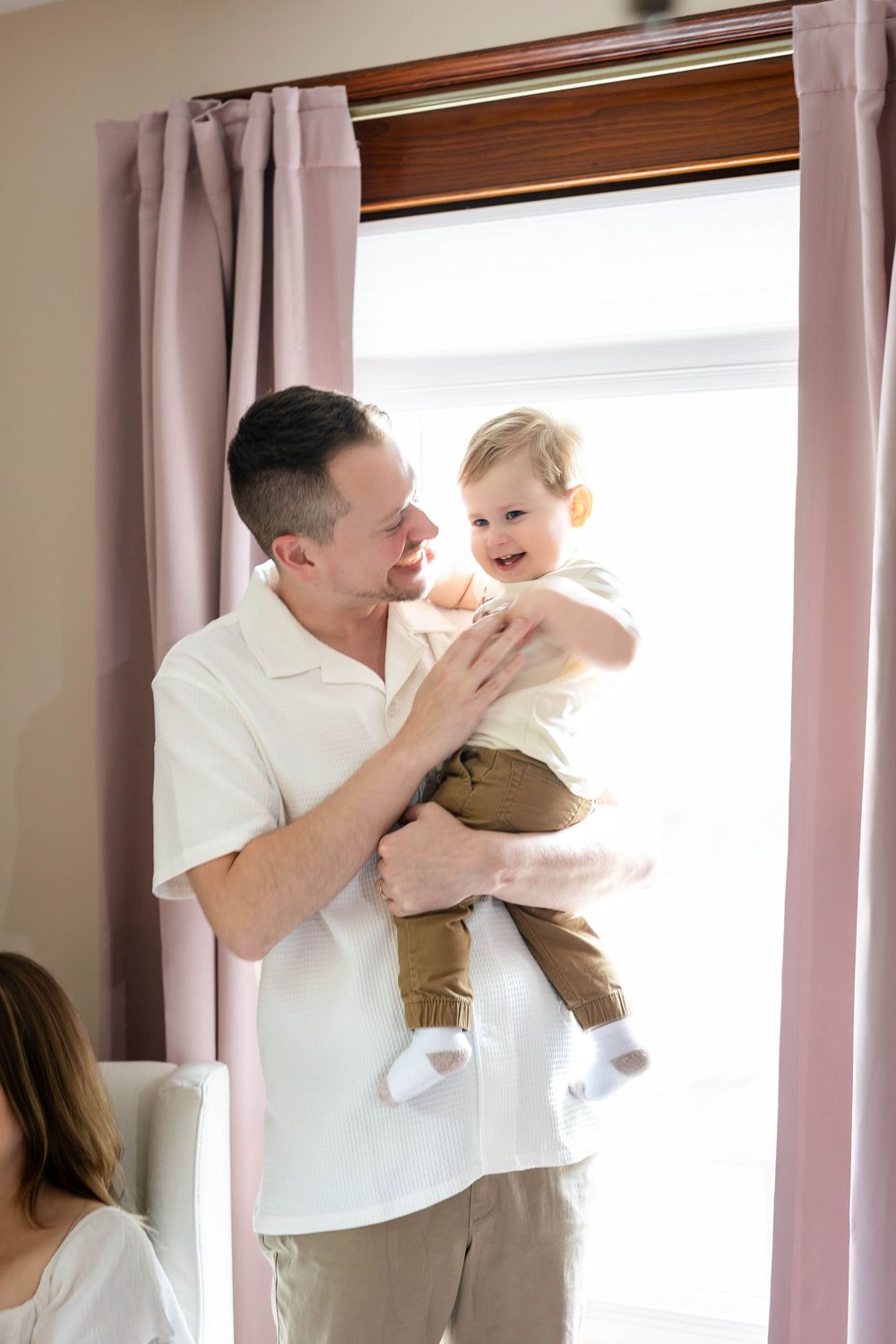 A joyful moment as a father in white lifts his smiling toddler by a window with pink curtains.
