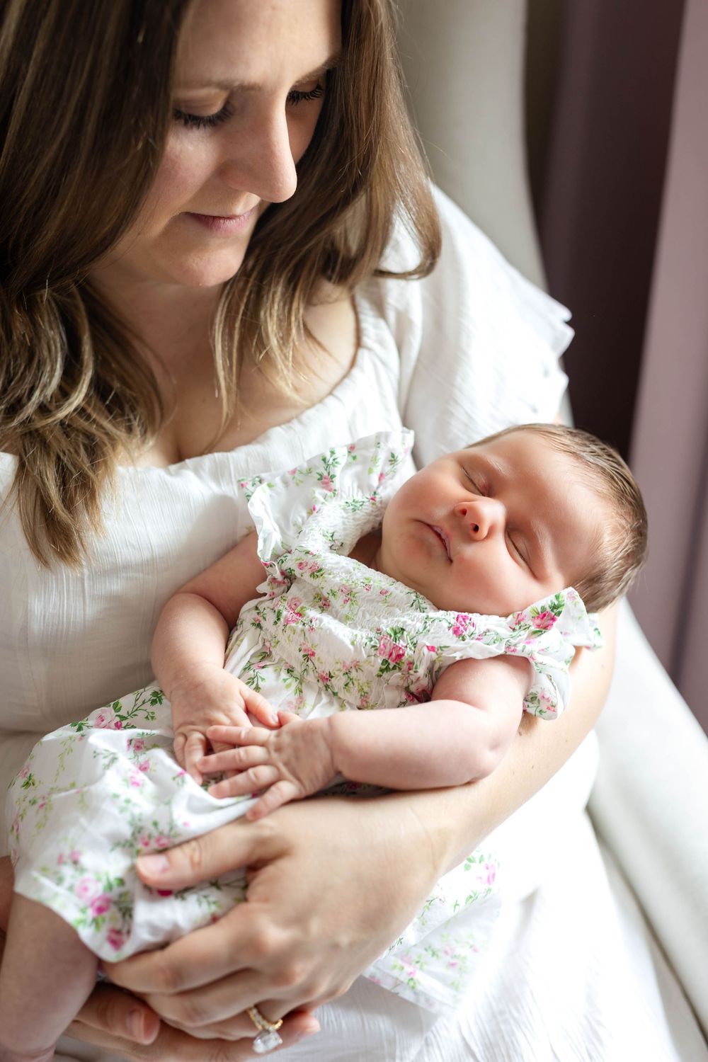 Tender moment of parent cradling sleeping infant wearing floral dress near window.
