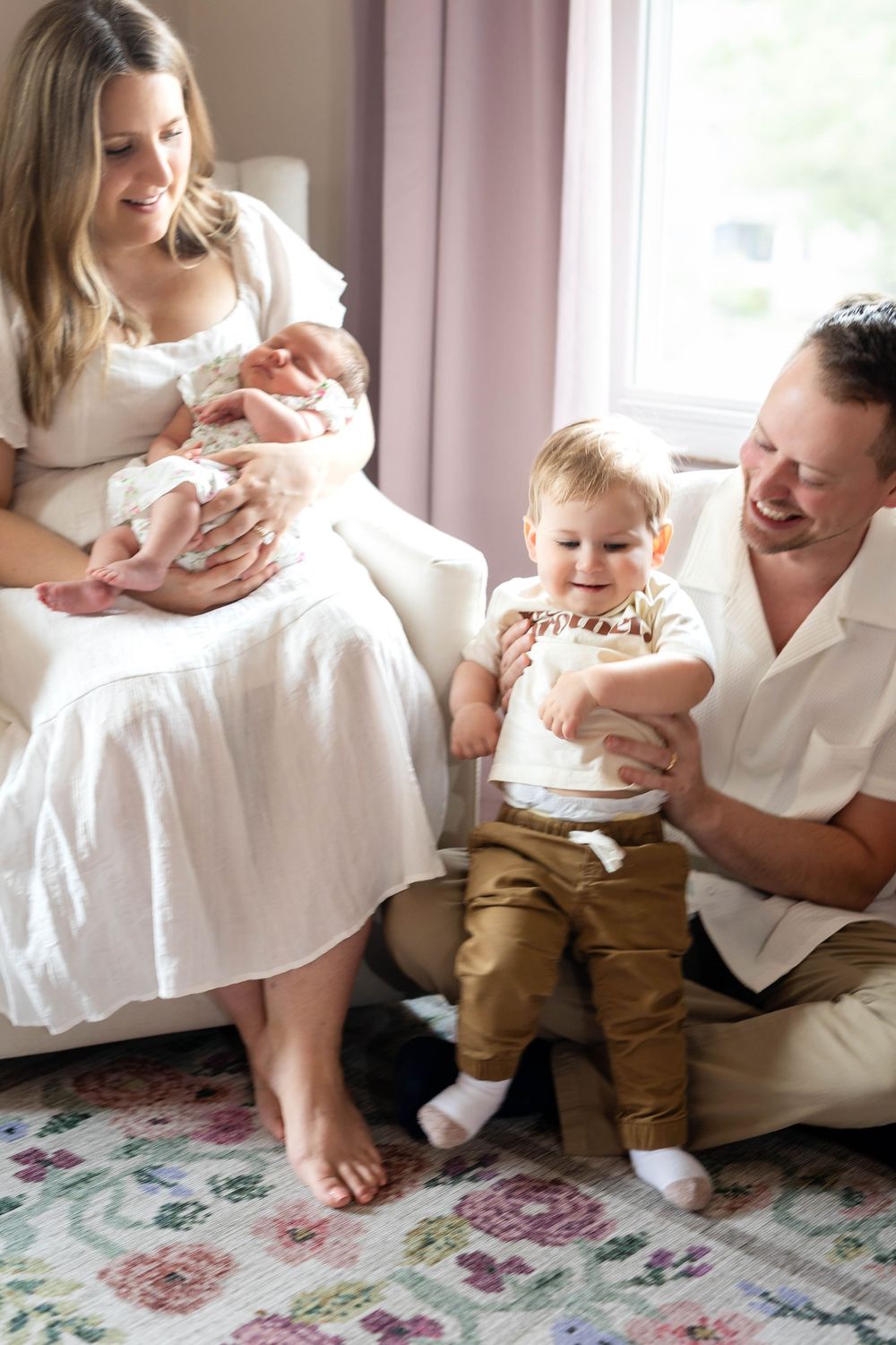 Family sitting together on floor by window with newborn baby and toddler.