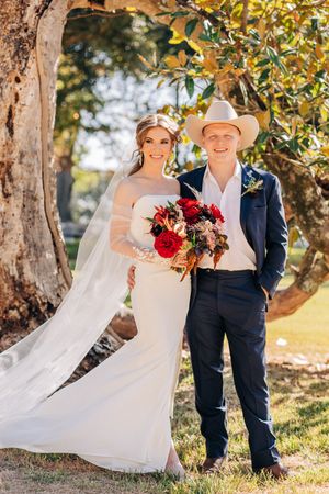 A couple in wedding attire poses beneath autumn trees with a flowing white veil and red bouquet.