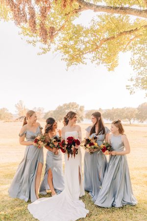 Bridesmaids in flowing dusty blue dresses gather with bride holding vibrant fall bouquets under golden autumn trees.
