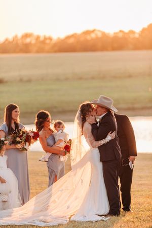 Wedding party gathered at sunset by a lake with bridesmaids in gray dresses and flowing bridal veil in golden light.